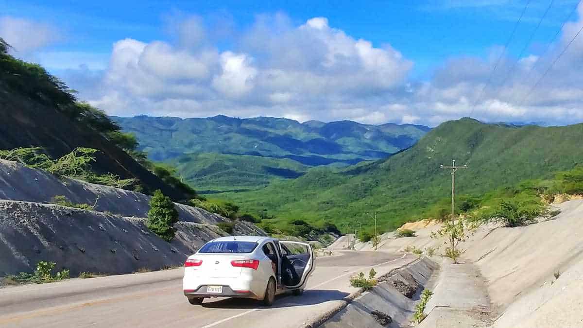 a white car is parked on the side of the road driving in dominican republic. in the distance there are rolling green hills.