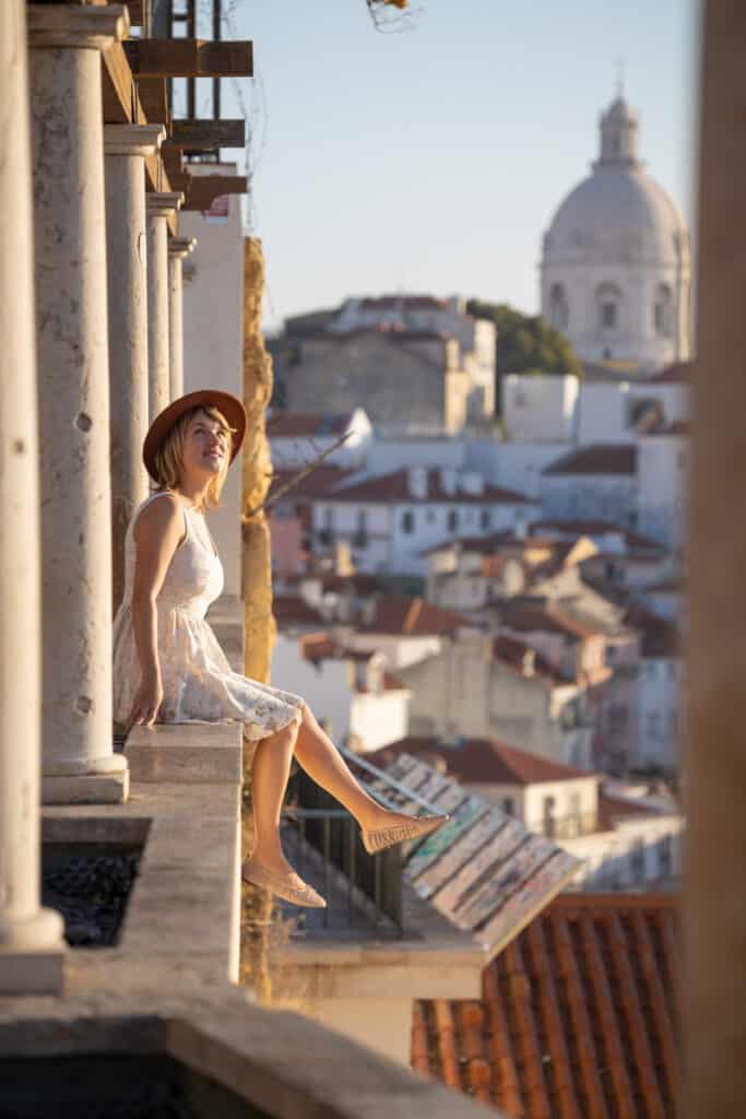 lora sitting on ledge in lisbon portugal. the background of the city is behind her and she is smiling looking out into the distance