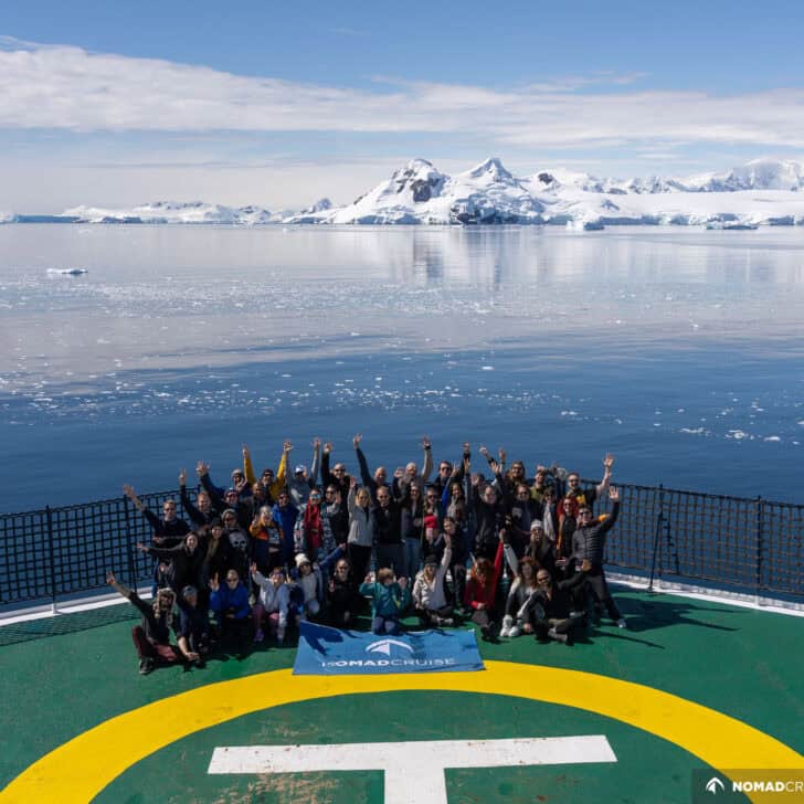A group of people cheering on a ship's helipad with serene icy waters and snow-covered mountains in the background.