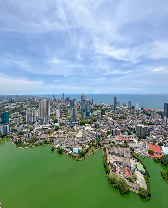 Aerial view of a cityscape with numerous high-rise buildings, overlooking a green-colored lake and the ocean in the background beneath a partly cloudy sky.