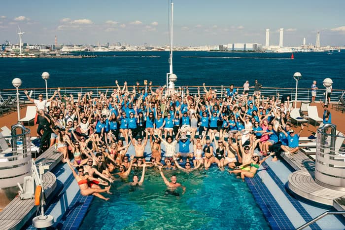 A lively crowd in blue shirts poses enthusiastically on a ship deck by a pool, with a city skyline and ocean backdrop, capturing the joyful spirit of their nomad cruise adventure.