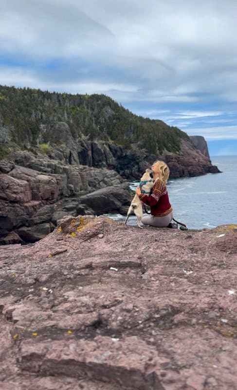 Person sitting on a rocky cliff, holding a dog and overlooking a vast expanse of ocean with rugged cliffs in the background under a cloudy sky.