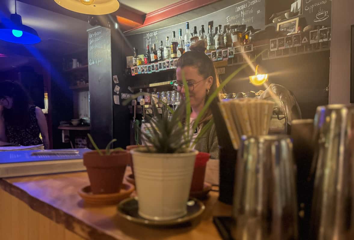 A person stands behind a bar counter with potted plants in the foreground and a variety of bottles and photos displayed on shelves in the background.