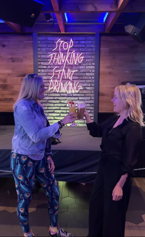 Two women clink glasses in front of a neon sign reading 