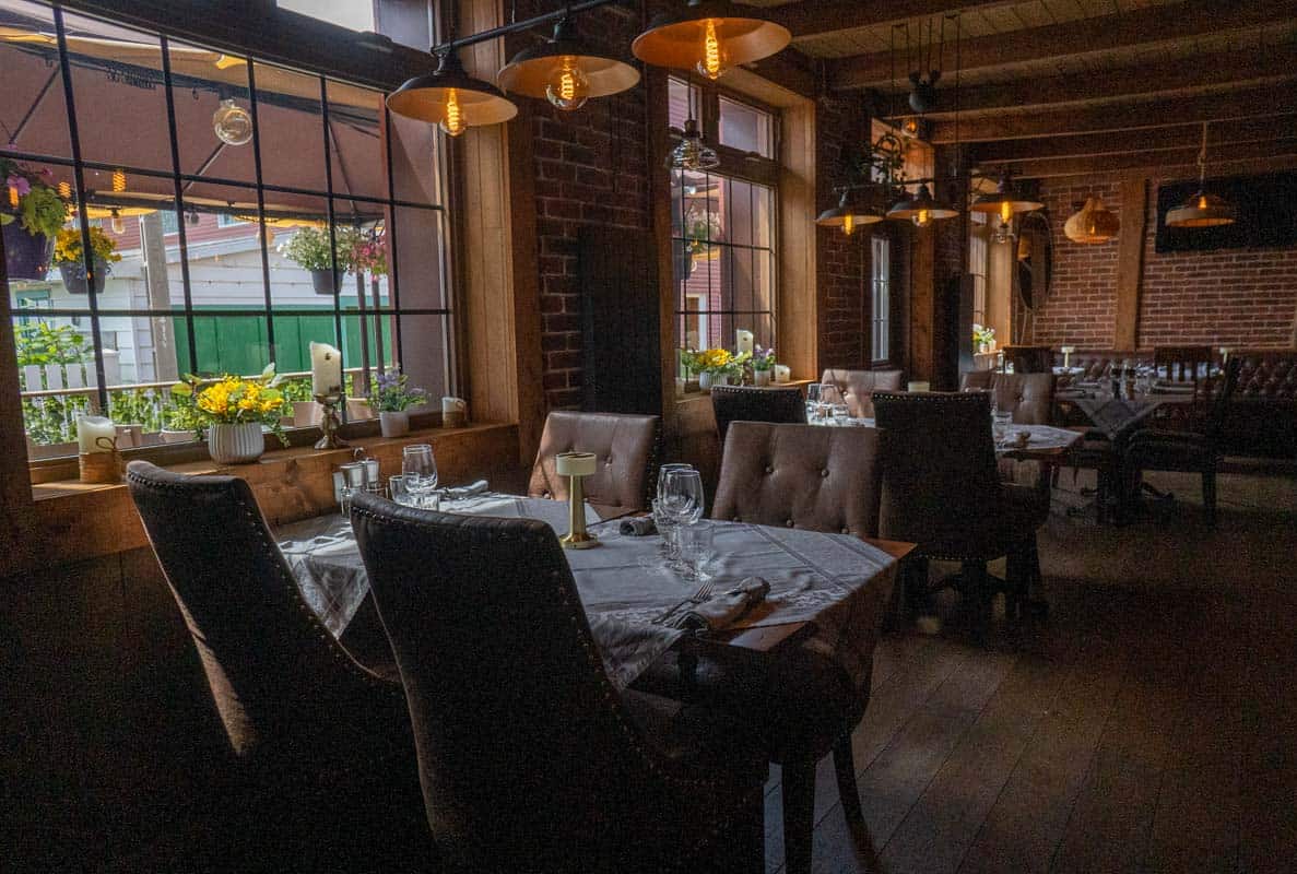 A cozy restaurant interior with wooden floors, brick walls, and large windows letting in natural light. Tables are set with white tablecloths, glassware, and yellow flowers.