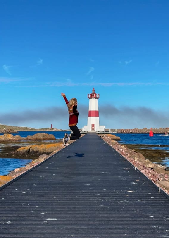 Person jumps on a pier with a red and white lighthouse in the background, clear blue sky above.