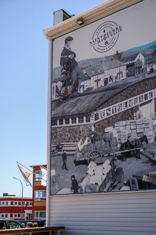 A large mural on the side of a building depicts a historical scene with people working and transporting goods. A modern street and colorful buildings are visible in the background.