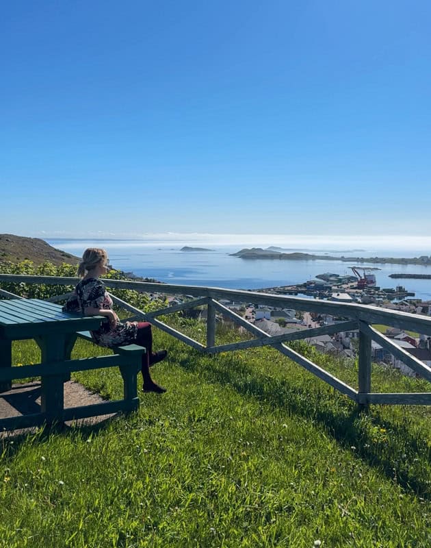 A person sits at a picnic table on a grassy hill overlooking a coastal town and the ocean on a clear, sunny day.