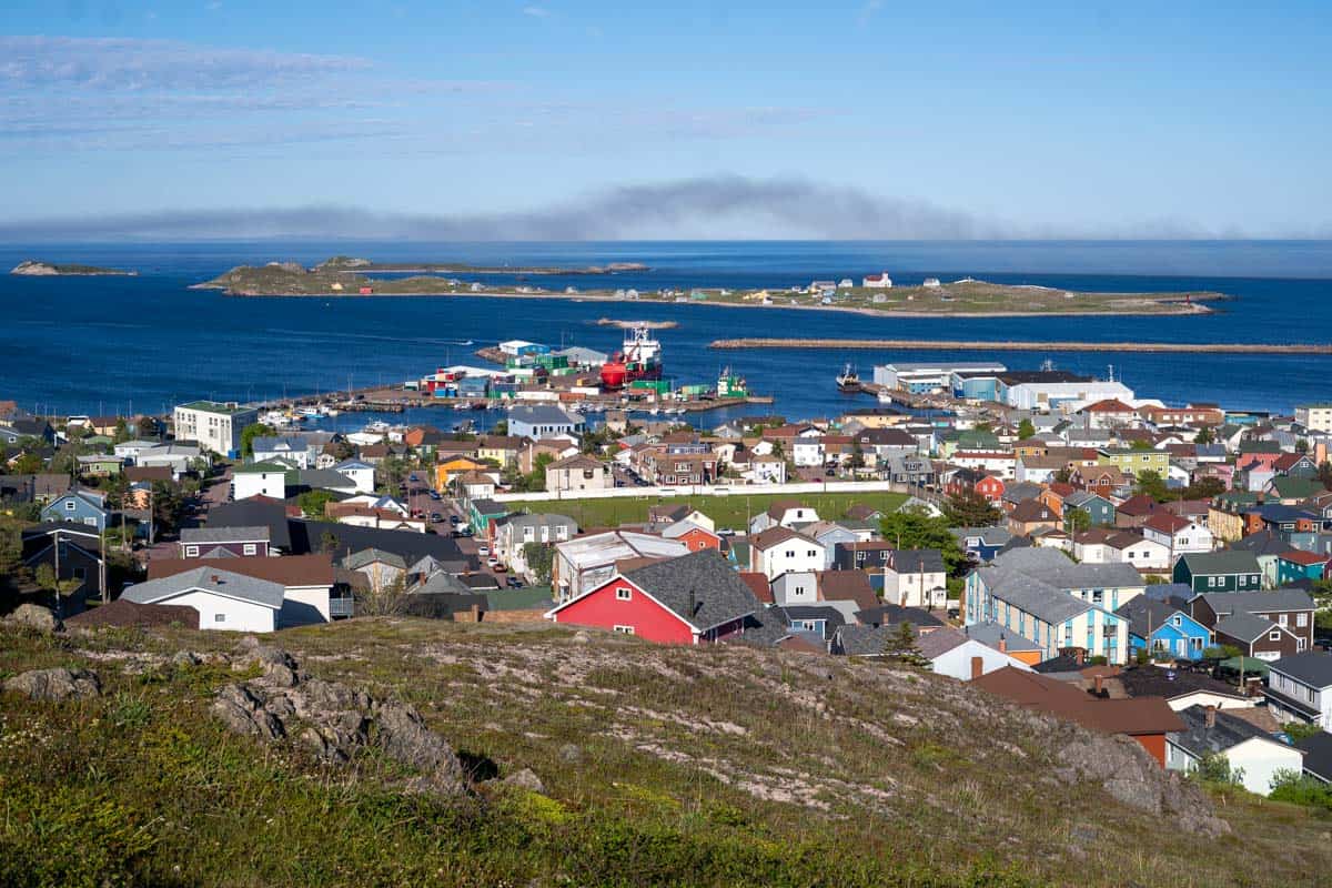 Aerial view of a coastal town with colorful houses, a harbor with boats, and islands in the background under a clear blue sky.