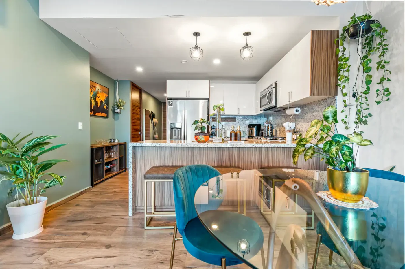 Modern kitchen with white cabinets, stainless steel appliances, and a marble island. The dining area features a glass table and blue chairs, with various plants adding greenery to the space.