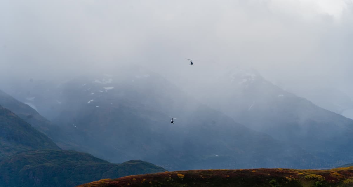 Two helicopters flying over misty mountains with rugged terrain and cloudy sky.