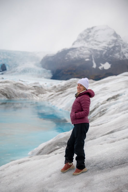 A person wearing a purple jacket and white beanie stands on snowy terrain near a blue lake, with a glacier and mountain in the background.