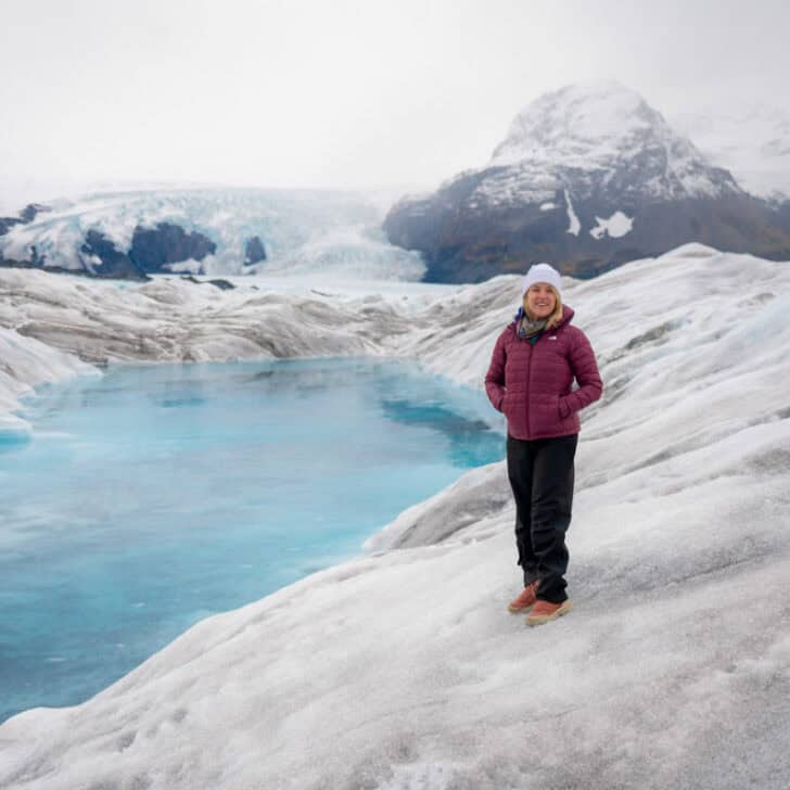 A person in a red jacket stands on a glacier near a blue glacial pool with snowy mountains in the background.