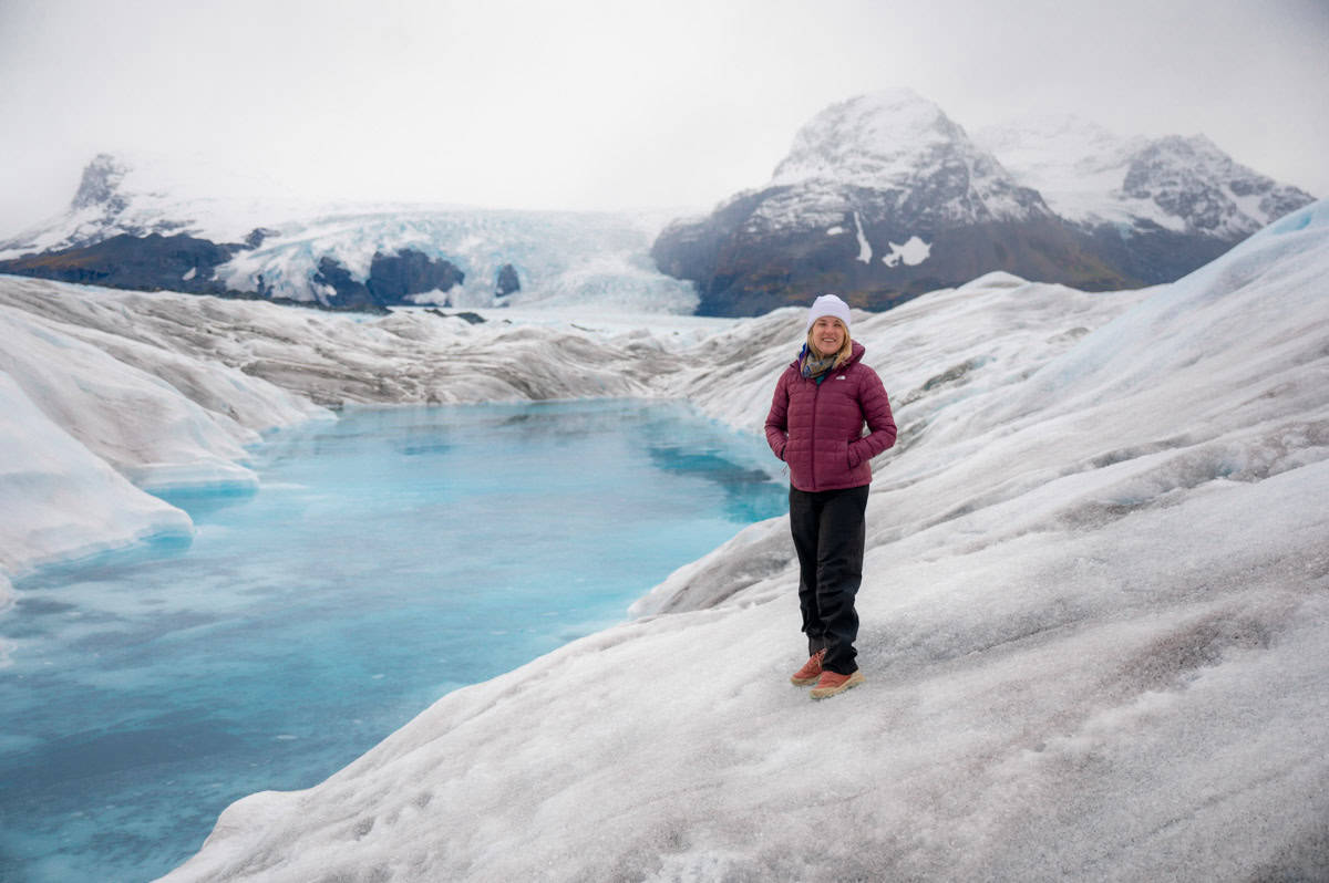 A person in a red jacket stands on a glacier near a blue glacial pool with snowy mountains in the background.