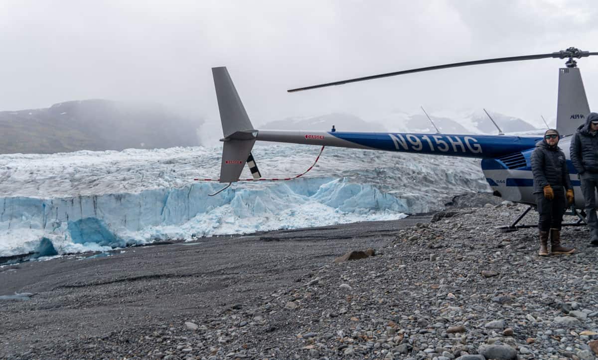 A helicopter is parked on rocky ground near a large glacier. Two people in winter clothing stand beside it. Cloudy sky and mountains are in the background.