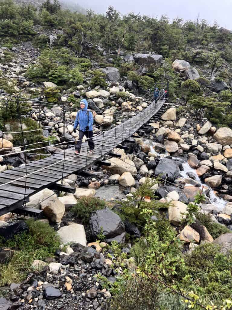 A person crosses a narrow suspension bridge over a rocky stream in a mountainous, misty landscape with greenery.