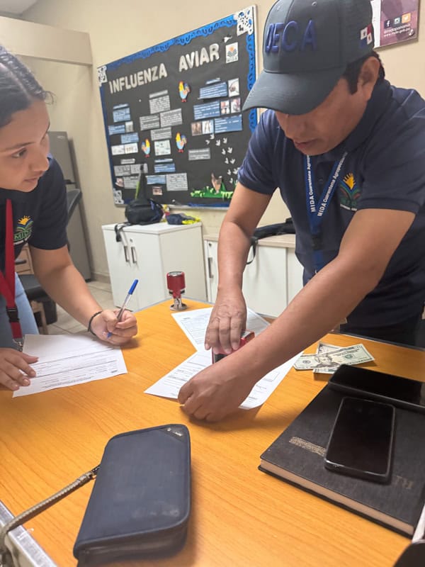 Two people in uniforms are working at a desk with papers, a stamp, a pen, money, and a phone. A wall with informational posters is in the background.