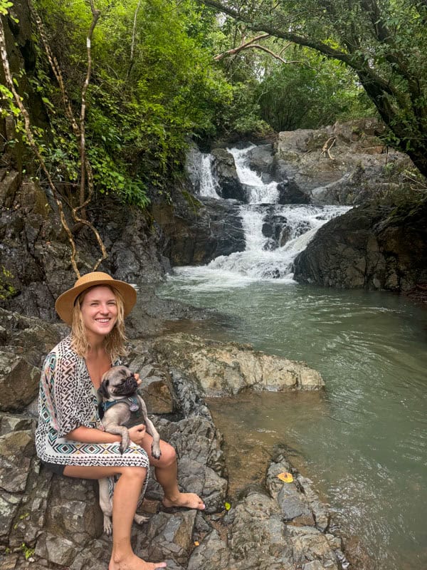 Woman in a hat sits on rocks by a waterfall, holding a small dog. Surrounded by lush greenery.