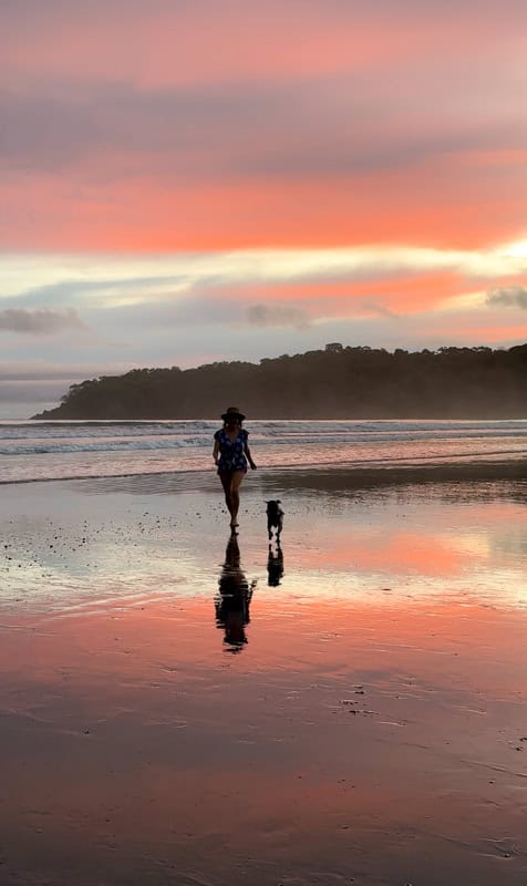 Person walking a dog on a reflective wet beach at sunset with a colorful sky and silhouetted hills in the background.