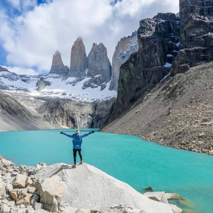 Person standing on a rock with arms raised, overlooking a turquoise lake and rugged mountain peaks under a partly cloudy sky.