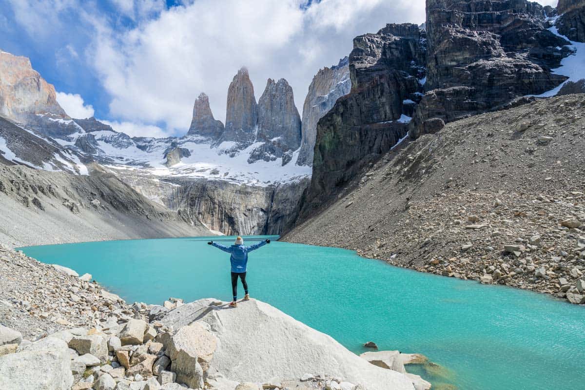 Person standing on a rock with arms raised, overlooking a turquoise lake and rugged mountain peaks under a partly cloudy sky.