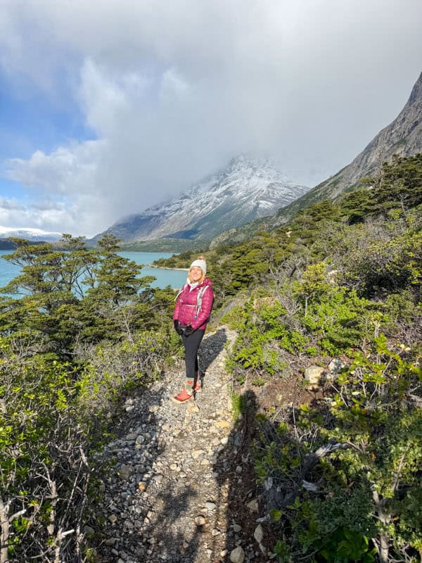 A person in a pink jacket stands on a rocky path surrounded by greenery, with a mountain and cloudy sky in the background.