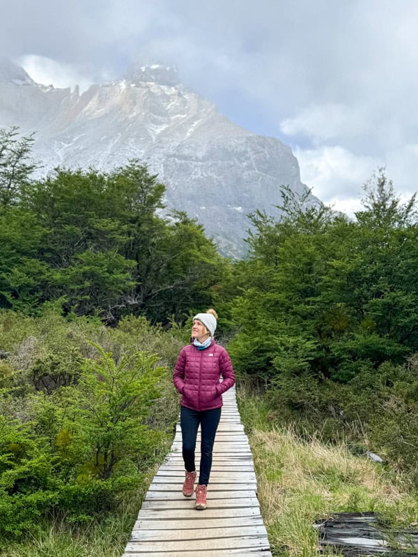 A person in a purple jacket walks on a wooden path surrounded by green vegetation, with mountains visible in the background.