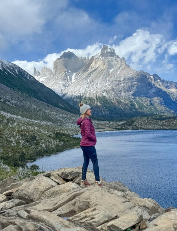 Person standing on rocks by a lake, wearing a red jacket and a beanie, with mountains in the background under a cloudy sky.