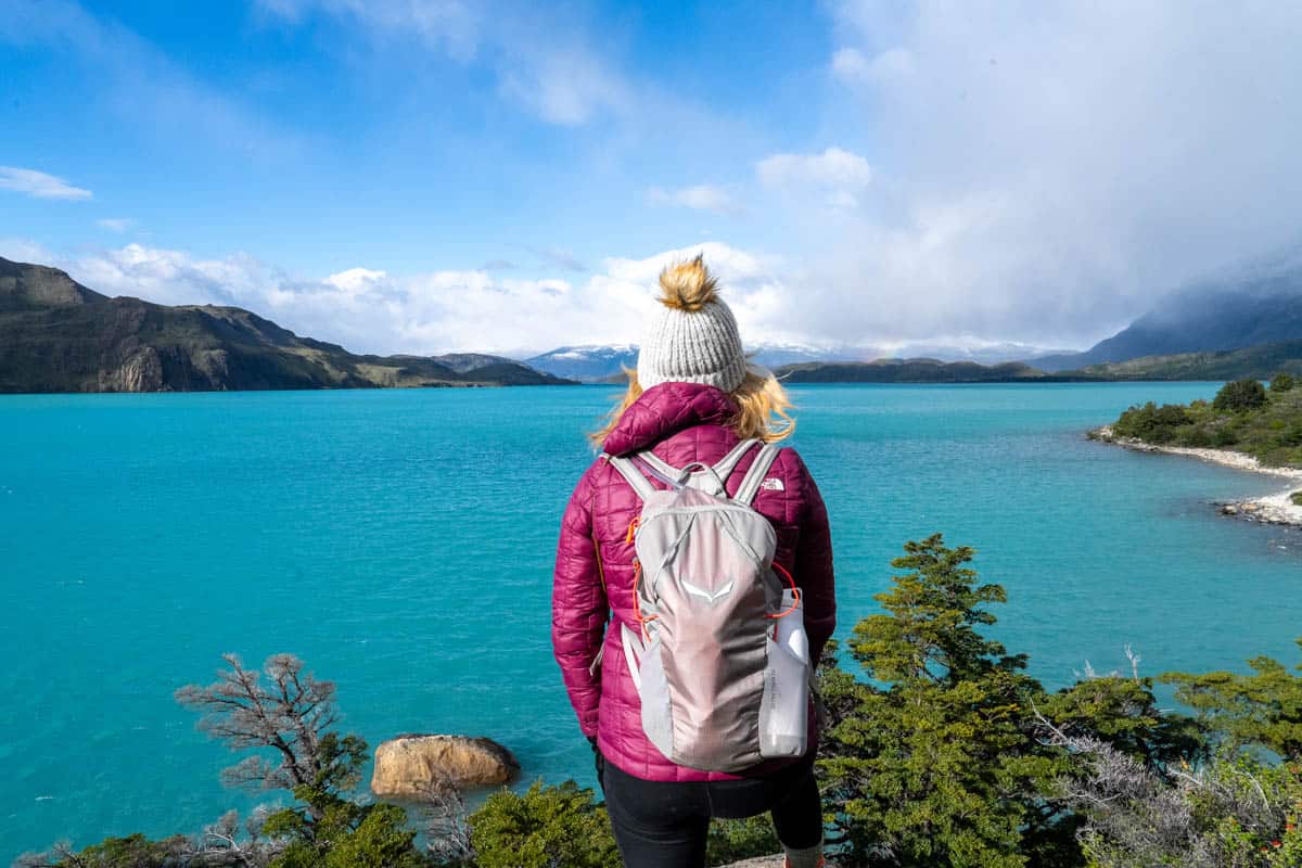 Person in a pink jacket and grey beanie stands facing a turquoise lake, with mountainous terrain and a partly cloudy sky in the background.