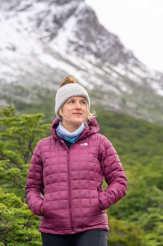 Person wearing a pink jacket and gray beanie stands in front of a green forest and snow-covered mountain.