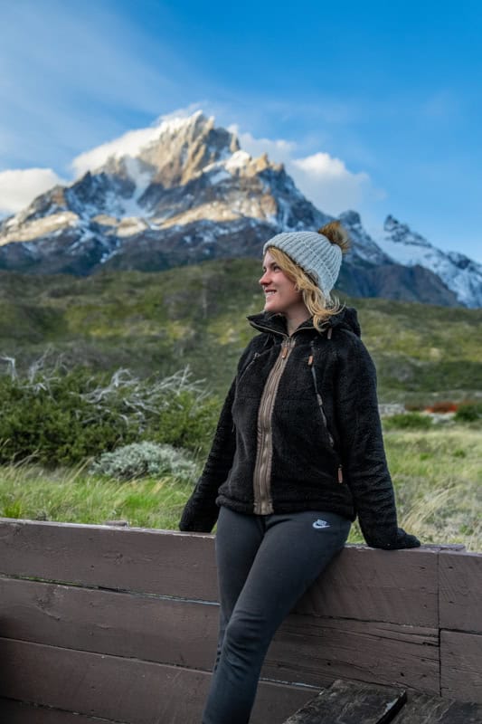 A person in a black jacket and beanie leans against a wooden fence, with a mountain and clear blue sky in the background.