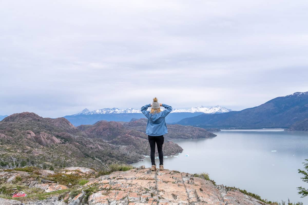 Person in a blue jacket and beanie stands on a rocky cliff overlooking a vast lake with distant snow-capped mountains.