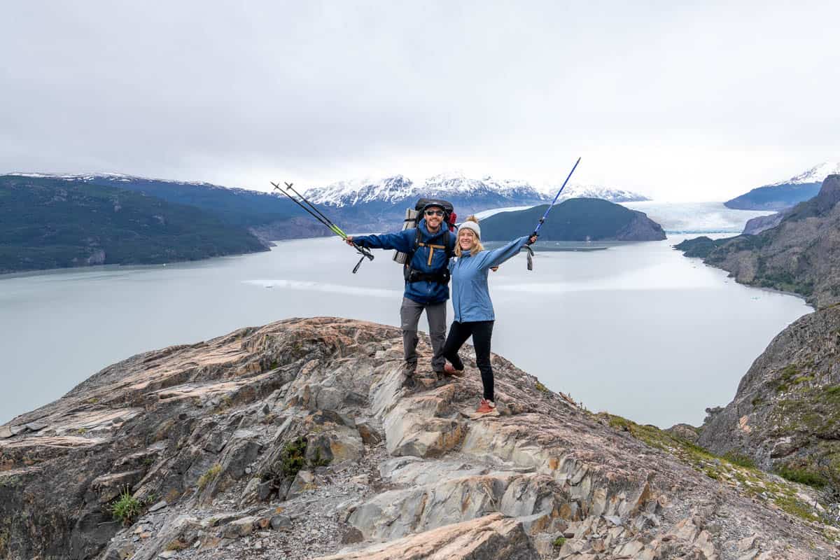 Two people standing on a rocky cliff, posing with trekking poles, overlooking a lake and snow-capped mountains.