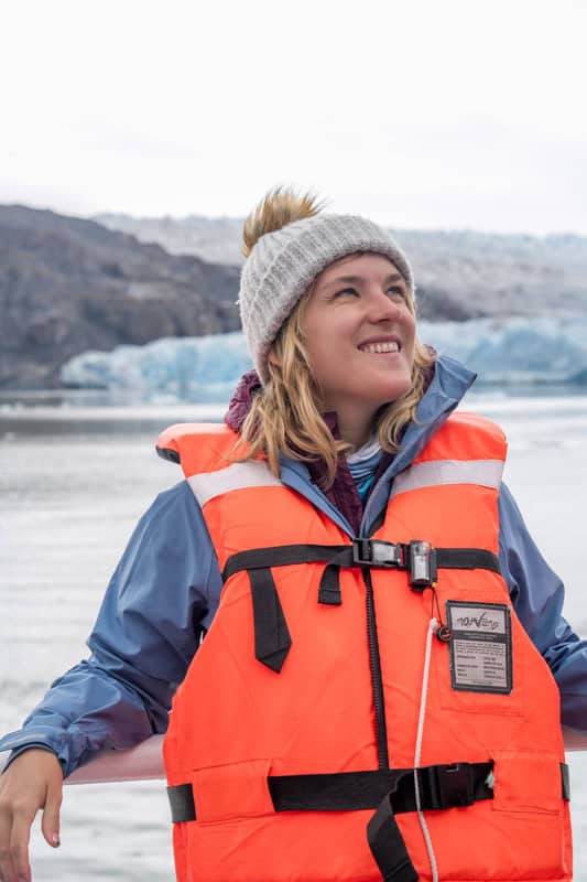 Person wearing a life jacket and beanie smiles while standing in front of a glacier.