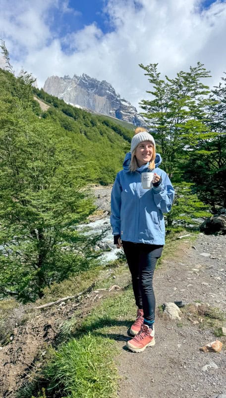 A person in a blue jacket and beanie holds a mug while standing on a trail in a lush mountainous landscape.