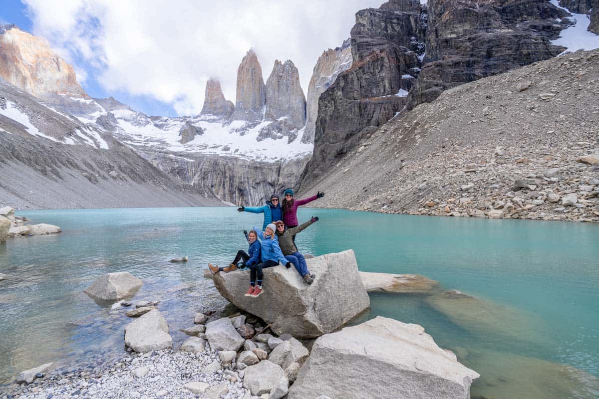 Four people sit on a rock by a turquoise lake with snow-capped mountains in the background. They are smiling and have their arms raised.